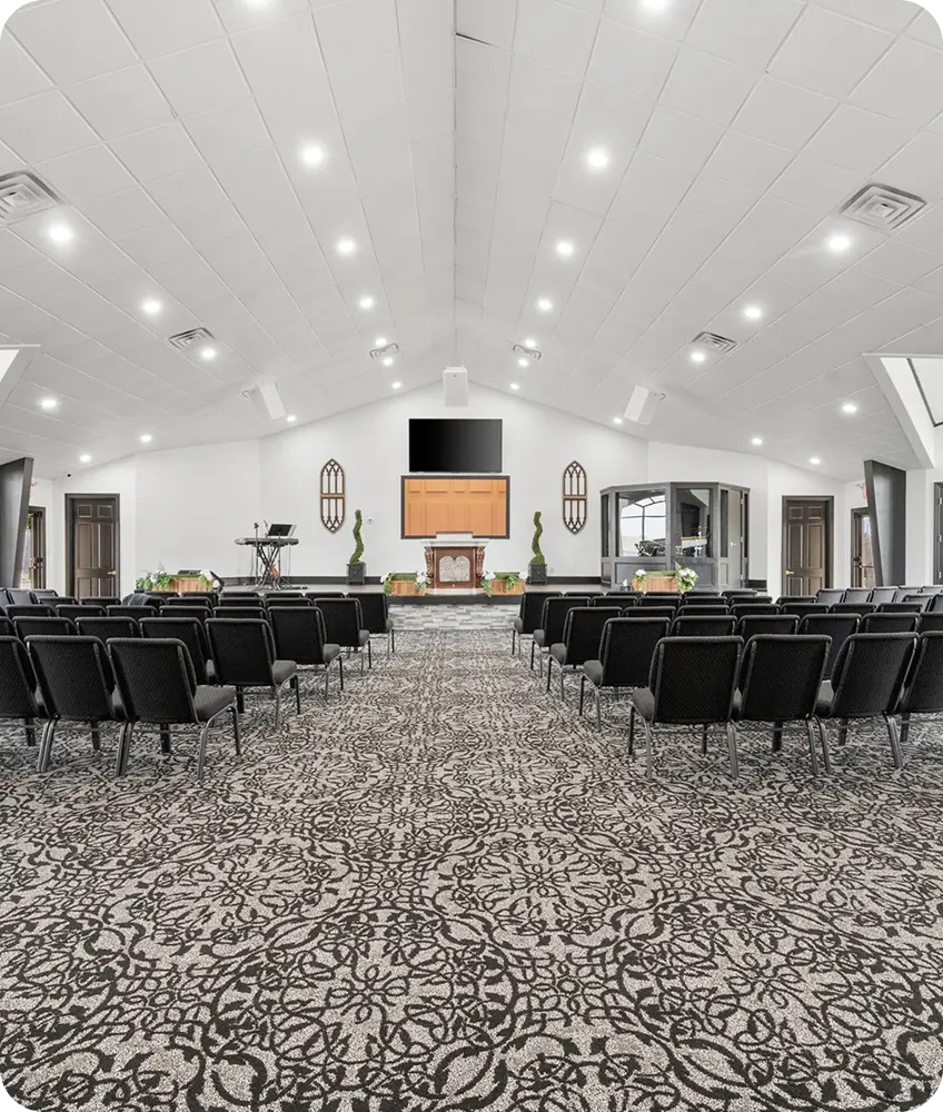 Interior view of a modern church sanctuary featuring rows of black chairs, a patterned carpet, a central altar, and vaulted ceilings with recessed lighting.