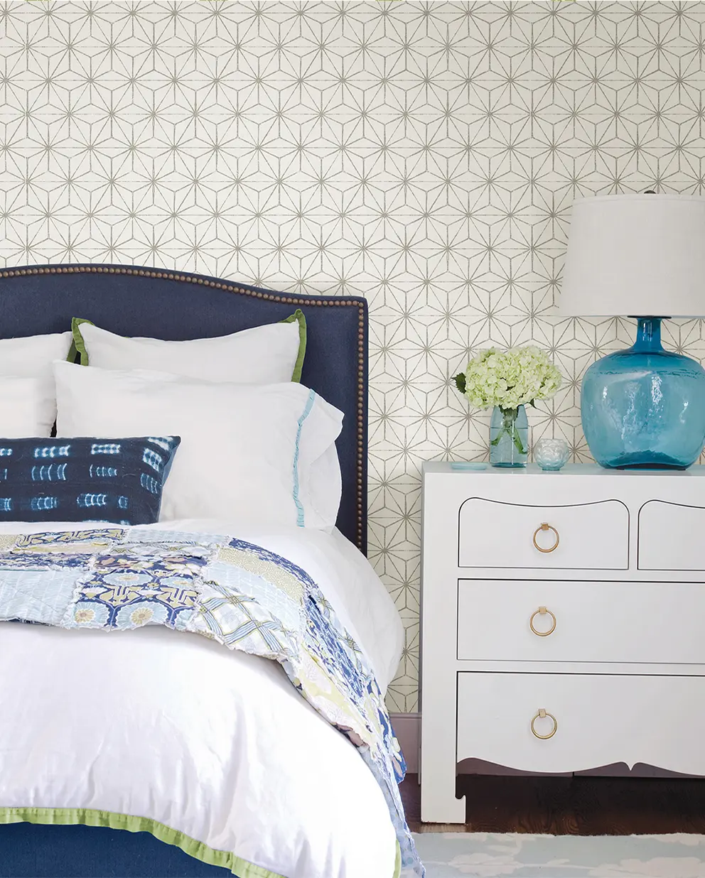 A bedroom scene highlighting a white wallpaper with a delicate, repeating geometric starburst pattern behind a blue headboard and white nightstand.